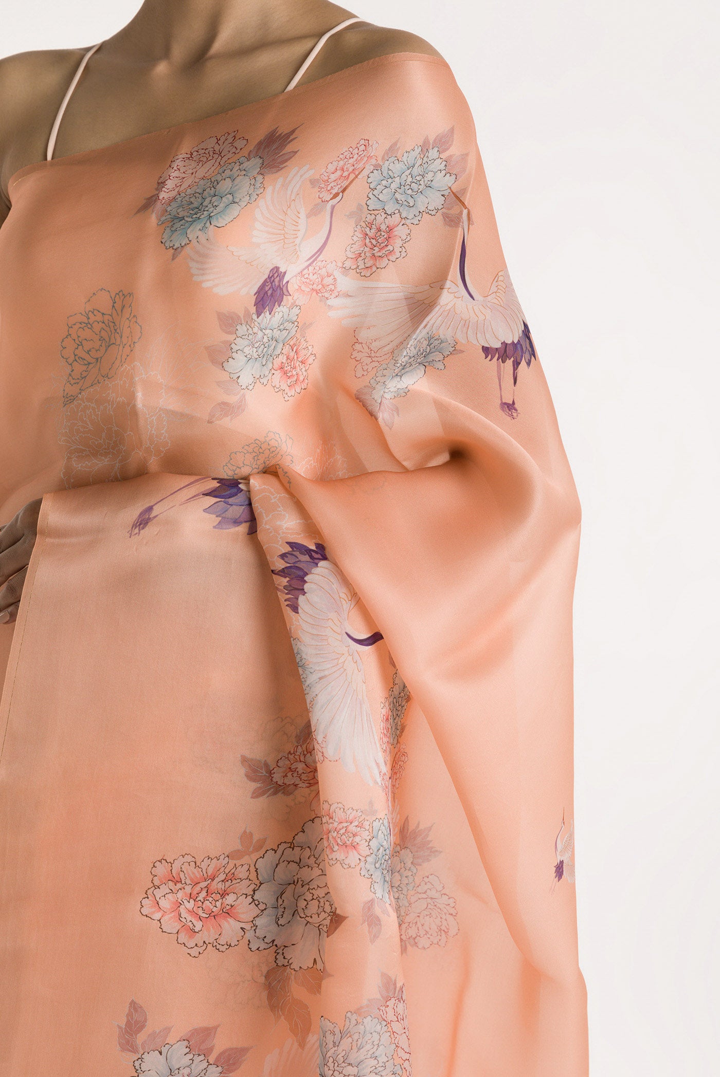 Close up of Our 'Flying Cranes' printed on a satin organza saree is crafted in a peach tone that features hand-illustrated tropical prints of flying cranes in the endless sky. The saree is finished with an intricately detailed thin Zari line along the border. displaying folds