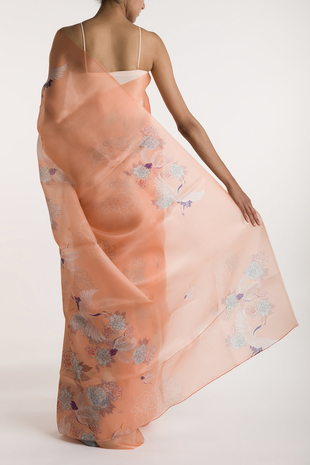 Pleats and pallu of Our 'Flying Cranes' printed on a satin organza saree is crafted in a peach tone that features hand-illustrated tropical prints of flying cranes in the endless sky. The saree is finished with an intricately detailed thin Zari line along the border.
