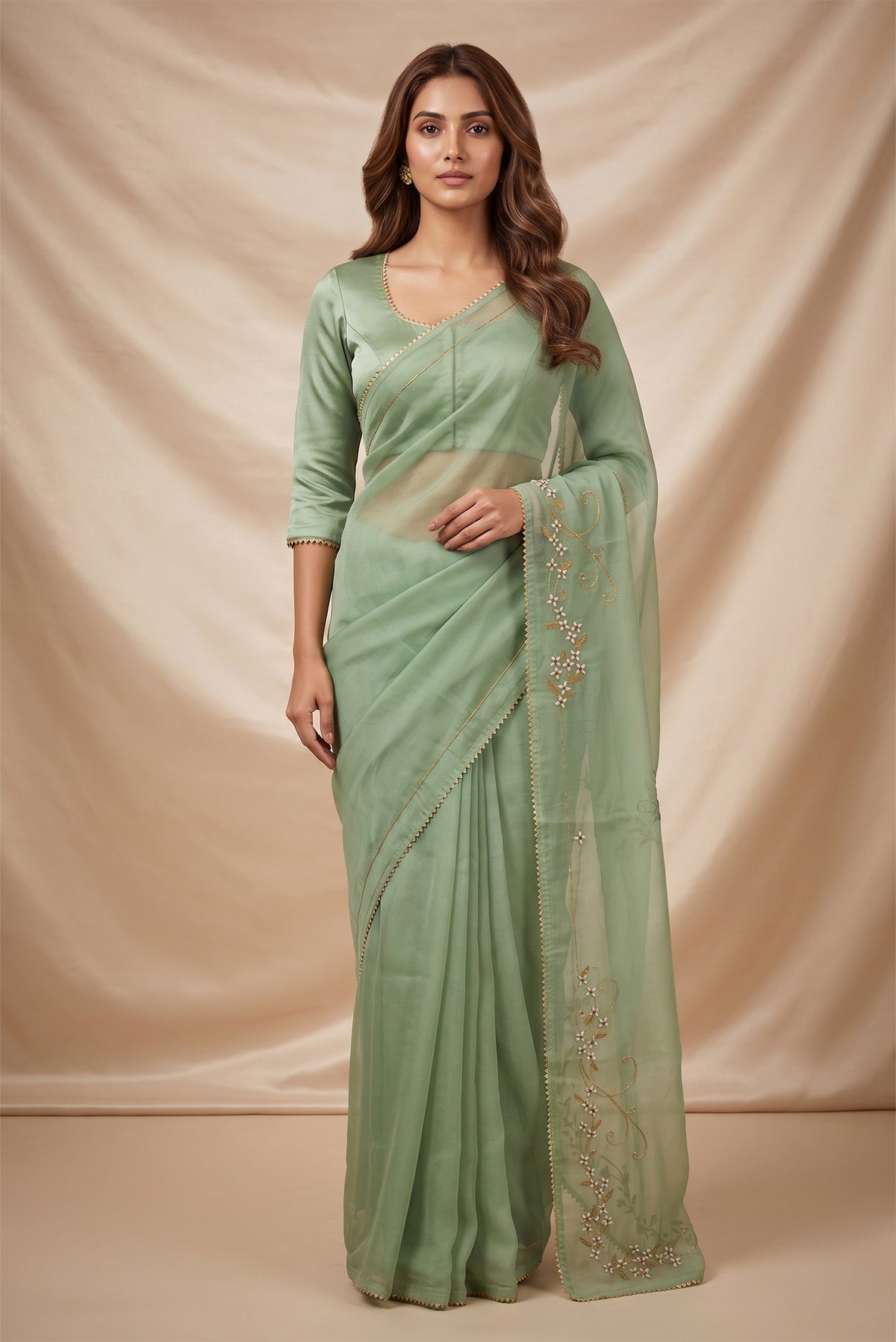 Model wearing a Mahira organza saree is inspired by the hydrangea blooms of the Art Nouveau movement. Along the border and edges, bold, simplified hydrangea blooms pair with fluid, curving stems. The flowers gain soft dimension through pani-zari zardozi, while antique-gold aari embroidery adds depth and balance. The pallu features three large asymmetrical butas, formed as flowing clusters of hydrangea blooms and stems, creating a graceful focal point.