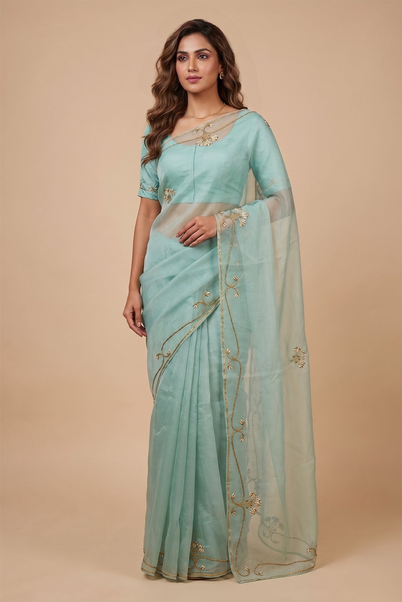 Model wearing a Meher, crafted in silk organza, is hand-embroidered with gold and antique-gold zardozi. Organic vines and floral buds form a rich border that meets at a delicate fan motif - an ornamental form celebrated in late 19th and early 20th century Art Nouveau design. Iris flowers, symbolising femininity, grace, and mystique, echo the saree?s fluid curves as the motif repeats across the body, finished with elegant gota edges.